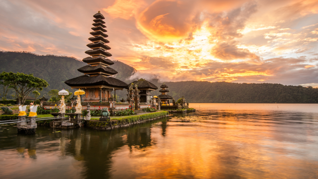 Sunset view of the Pura Ulun Danu Bratan temple on Lake Bratan in Bali, Indonesia, with a serene reflection of the temple and vibrant orange sky on the calm water, surrounded by lush green mountains.