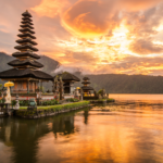 Sunset view of the Pura Ulun Danu Bratan temple on Lake Bratan in Bali, Indonesia, with a serene reflection of the temple and vibrant orange sky on the calm water, surrounded by lush green mountains.