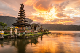 Sunset view of the Pura Ulun Danu Bratan temple on Lake Bratan in Bali, Indonesia, with a serene reflection of the temple and vibrant orange sky on the calm water, surrounded by lush green mountains.