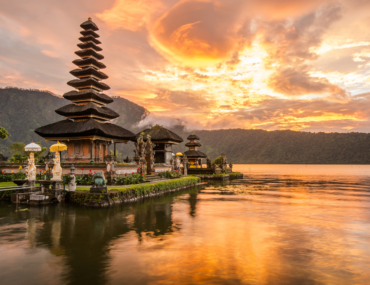 Sunset view of the Pura Ulun Danu Bratan temple on Lake Bratan in Bali, Indonesia, with a serene reflection of the temple and vibrant orange sky on the calm water, surrounded by lush green mountains.