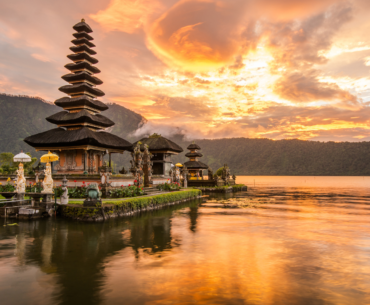 Sunset view of the Pura Ulun Danu Bratan temple on Lake Bratan in Bali, Indonesia, with a serene reflection of the temple and vibrant orange sky on the calm water, surrounded by lush green mountains.