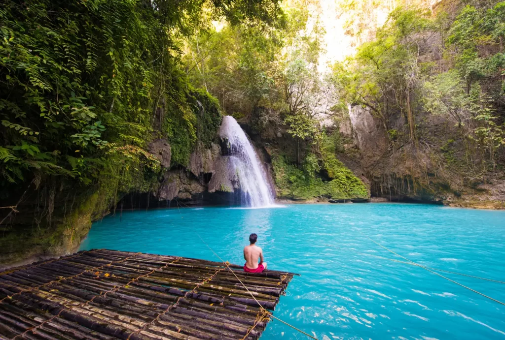  A man relaxes on a bamboo raft in turquoise water, with the stunning Kawasan Falls cascading in the background.