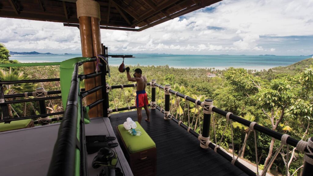 Muay Thai training ring with a scenic ocean view, surrounded by lush greenery, located at an elevated platform at Four Seasons Resort Koh Samui. A man in red and blue boxing shorts practices with a speed bag under a traditional wooden pavilion.