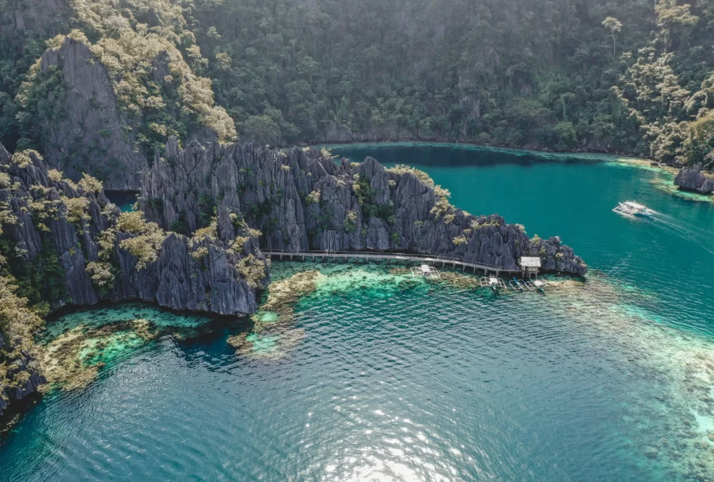 An aerial view of a stunning tropical destination with dramatic limestone cliffs covered in lush greenery, turquoise waters, and coral reefs visible below the surface. A wooden boardwalk stretches along the base of the cliffs, leading to small boats docked near the shore. In the distance, a motorized boat glides across the pristine water, completing the picturesque scene.