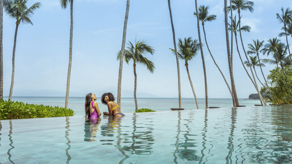 Two women enjoying a sunny day in a pool, surrounded by lush palm trees in the background.