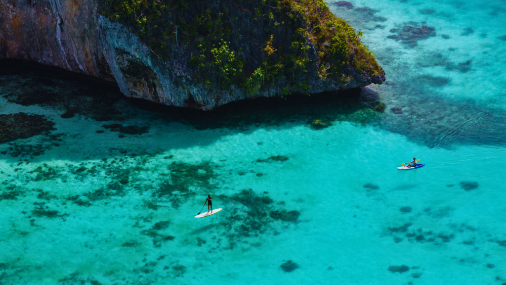 "Two travelers enjoying water activities in the crystal-clear turquoise waters of Raja Ampat, surrounded by lush limestone cliffs and vibrant coral reefs, showcasing the natural beauty of this Indonesian paradise."