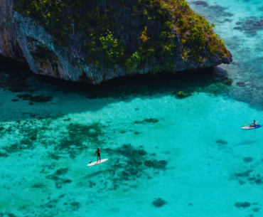 "Two travelers enjoying water activities in the crystal-clear turquoise waters of Raja Ampat, surrounded by lush limestone cliffs and vibrant coral reefs, showcasing the natural beauty of this Indonesian paradise."