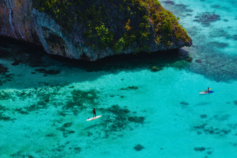 "Two travelers enjoying water activities in the crystal-clear turquoise waters of Raja Ampat, surrounded by lush limestone cliffs and vibrant coral reefs, showcasing the natural beauty of this Indonesian paradise."