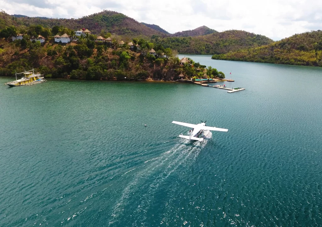 A seaplane preparing to take off or land on a calm, turquoise lake surrounded by lush, green hills. In the background, small houses or resorts are perched on the hillside, and a dock with boats is visible near the water's edge.