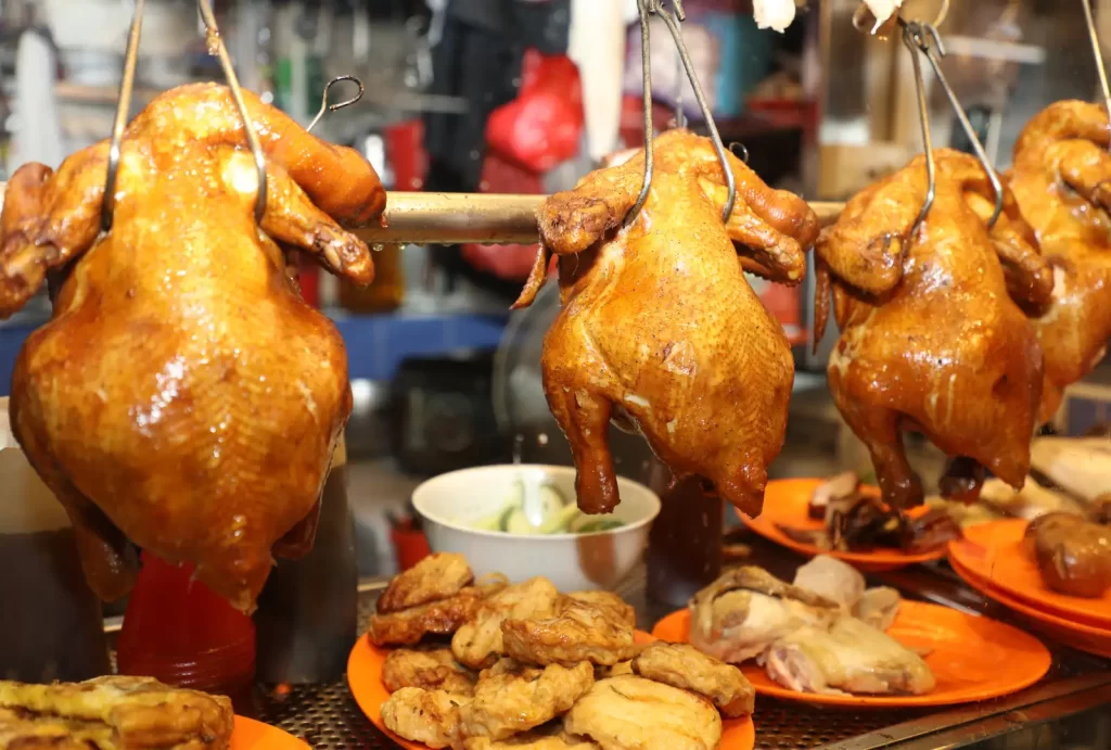 A display of roasted whole chickens hanging on hooks, accompanied by plates of sliced chicken and other side dishes, typical of Singapore's vibrant hawker stalls.