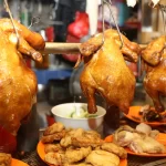 A display of roasted whole chickens hanging on hooks, accompanied by plates of sliced chicken and other side dishes, typical of Singapore's vibrant hawker stalls.