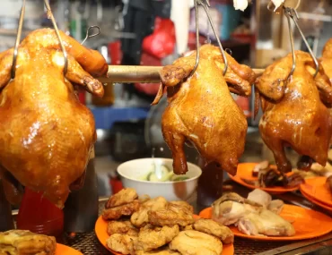 A display of roasted whole chickens hanging on hooks, accompanied by plates of sliced chicken and other side dishes, typical of Singapore's vibrant hawker stalls.