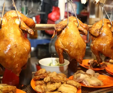 A display of roasted whole chickens hanging on hooks, accompanied by plates of sliced chicken and other side dishes, typical of Singapore's vibrant hawker stalls.