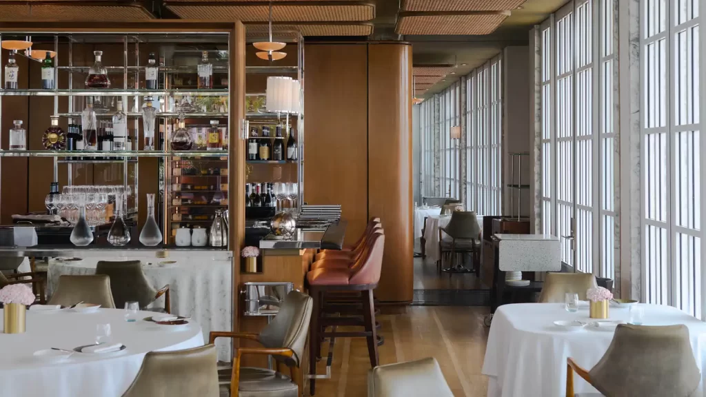 A luxurious and modern restaurant interior featuring elegant wooden panels, floor-to-ceiling windows with geometric designs, white tablecloth-covered tables, and a well-stocked bar area with leather stools.