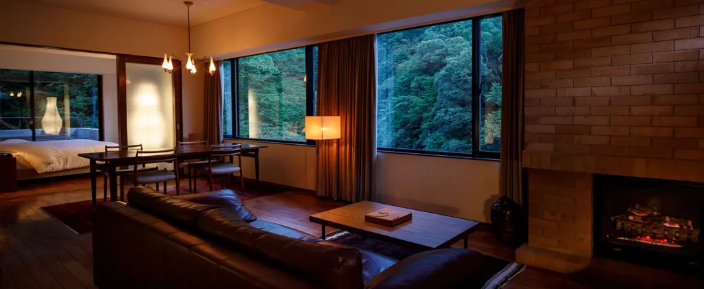 An elegant suite at Arcana Izu, featuring a living room with a fireplace and windows looking out into the serene forest. A unique, romantic stay for bespoke Japan travel and luxury Japan holidays.