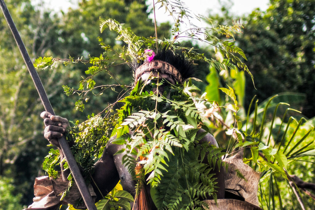 A man, representing an Asmat warrior, holds a spear while plants in his hair enhance his camouflage among the greenery.
