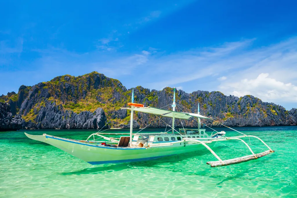 Traditional bangka outrigger boat in turquoise water beneath limestone cliffs, El Nido, Palawan