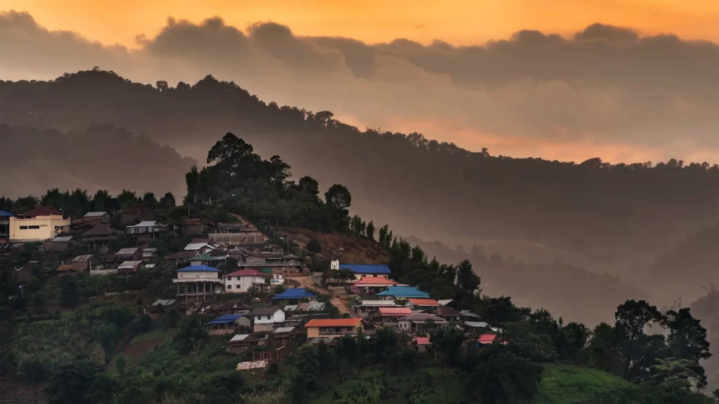 Hill tribe village perched on a misty hillside at sunset in Chiang Rai, northern Thailand, with layered mountain ridges fading into an amber sky