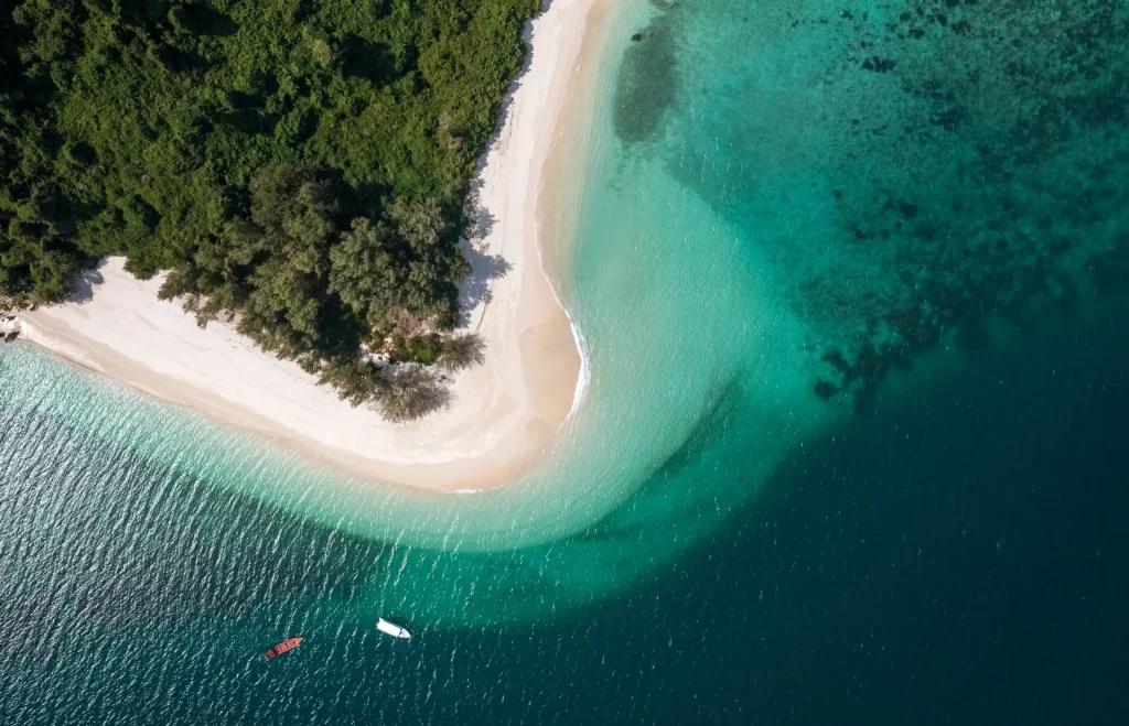 Aerial view of a pristine tropical island featuring a curved white sand beach, lush green jungle, and crystal-clear turquoise waters with two boats anchored offshore — Discovery DMC