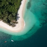 Aerial view of a pristine tropical island featuring a curved white sand beach, lush green jungle, and crystal-clear turquoise waters with two boats anchored offshore — Discovery DMC