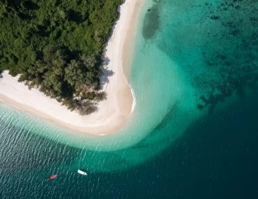 Aerial view of a pristine tropical island featuring a curved white sand beach, lush green jungle, and crystal-clear turquoise waters with two boats anchored offshore — Discovery DMC