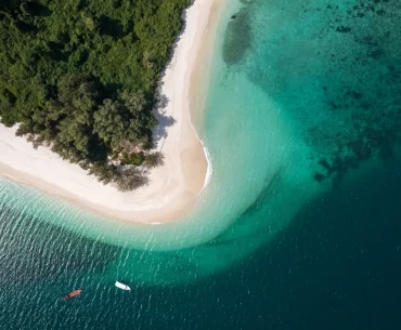 Aerial view of a pristine tropical island featuring a curved white sand beach, lush green jungle, and crystal-clear turquoise waters with two boats anchored offshore — Discovery DMC