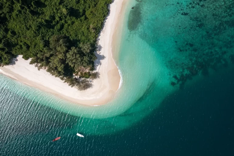 Aerial view of a pristine tropical island featuring a curved white sand beach, lush green jungle, and crystal-clear turquoise waters with two boats anchored offshore — Discovery DMC