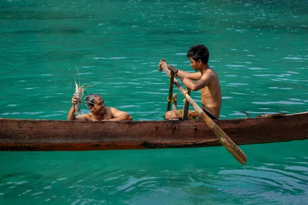 Moken sea nomad holding a freshly caught lobster in turquoise waters beside a traditional wooden canoe paddled by a young fisherman in the Andaman Sea .