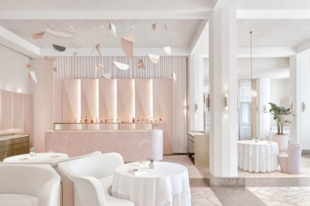 A fine-dining restaurant with a modern, minimalist aesthetic. The space is predominantly white and pastel pink, featuring elegant furnishings, round tables with white tablecloths, and plush chairs. The bar area is highlighted by a sleek, pink marble counter and an artistic, abstract installation of hanging shapes on the ceiling. The room is bright and airy, with natural light streaming through large windows and subtle decor details like potted plants and soft wall lighting.