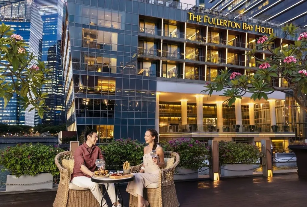 A smiling couple enjoying riverside outdoor dining and drinks in front of The Fullerton Bay Hotel, a perfect addition to luxury Singapore holidays and tours arranged by a Singapore tour operator.