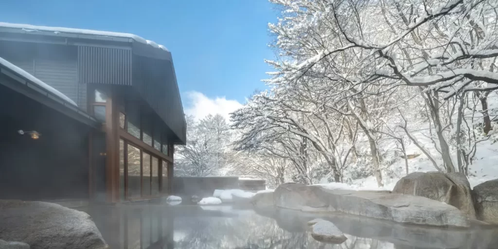 Steaming outdoor onsen at Hoshinoya Karuizawa in winter, surrounded by snow-laden trees — soaking in a natural hot spring while snow falls is one of Japan's defining luxury experiences