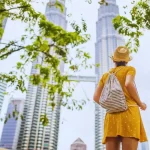 A traveler in a sunhat looks up at the iconic Petronas Twin Towers in Kuala Lumpur, the perfect destination for your next Malaysia holiday.