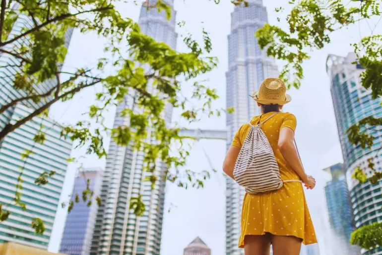 A traveler in a sunhat looks up at the iconic Petronas Twin Towers in Kuala Lumpur, the perfect destination for your next Malaysia holiday.