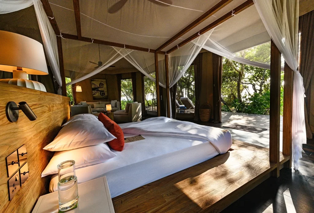 Interior of a luxury tented villa with a four-poster canopy bed dressed in white linen, red accent pillows, and open walls revealing a private deck and tropical jungle — Discovery DMC