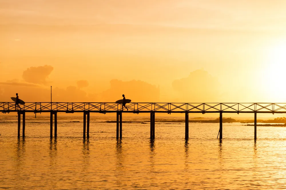 Silhouetted surfers carrying surfboards walk along a wooden boardwalk over the ocean during a golden sunset in Siargao, Philippines.
