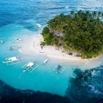 Aerial view of Guyam Island in Siargao, Philippines, featuring lush palm trees, white sand beaches, and traditional outrigger boats anchored in clear turquoise waters.