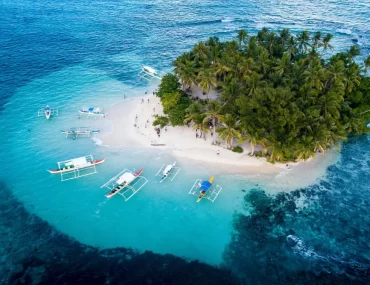 Aerial view of Guyam Island in Siargao, Philippines, featuring lush palm trees, white sand beaches, and traditional outrigger boats anchored in clear turquoise waters.