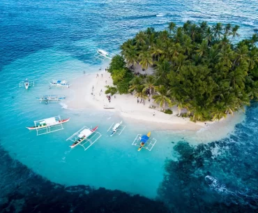Aerial view of Guyam Island in Siargao, Philippines, featuring lush palm trees, white sand beaches, and traditional outrigger boats anchored in clear turquoise waters.