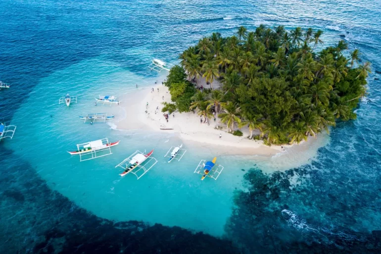 Aerial view of Guyam Island in Siargao, Philippines, featuring lush palm trees, white sand beaches, and traditional outrigger boats anchored in clear turquoise waters.