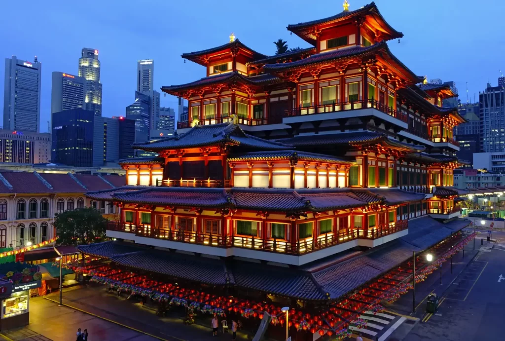 Stunning evening view of Singapore's Buddha Tooth Relic Temple in Chinatown, illuminated with warm lights, against a backdrop of modern skyscrapers.