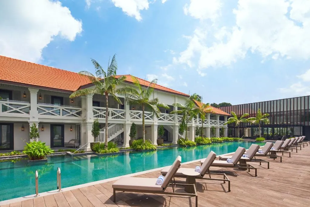 Stunning outdoor swimming pool at a luxury Singapore resort, featuring colonial architecture, wooden decking, palm trees, and modern sun loungers. A perfect feature for high-end Singapore travel and holidays.