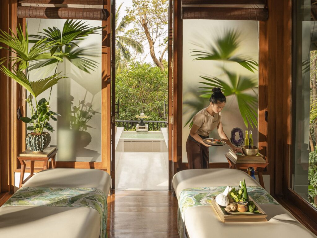 A tranquil spa room with tropical decor, featuring two massage tables adorned with botanical patterns, wooden furnishings, and vibrant green plants. A spa therapist prepares treatments in the natural sunlight streaming through open doors, with a lush outdoor garden view in the background.