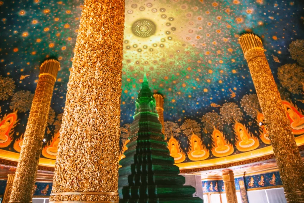 Ornate interior of a Thai Buddhist temple featuring intricate gold columns, a glowing emerald green chedi, and stunning celestial ceiling murals — cultural Thailand travel curated by a luxury Thailand DMC and tour operator