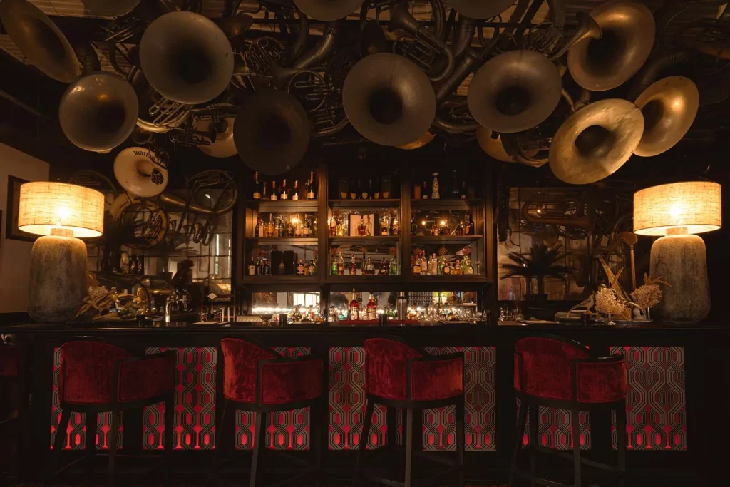 Interior of Thailand cocktail bar in Bangkok, with vintage brass instruments covering the ceiling above a backlit bar and red velvet stools — one of Bangkok's most distinctive luxury nightlife experiences