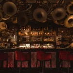 Interior of Thailand cocktail bar in Bangkok, with vintage brass instruments covering the ceiling above a backlit bar and red velvet stools — one of Bangkok's most distinctive luxury nightlife experiences