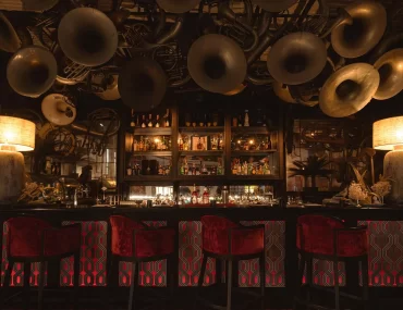 Interior of Thailand cocktail bar in Bangkok, with vintage brass instruments covering the ceiling above a backlit bar and red velvet stools — one of Bangkok's most distinctive luxury nightlife experiences