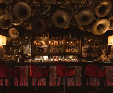Interior of Thailand cocktail bar in Bangkok, with vintage brass instruments covering the ceiling above a backlit bar and red velvet stools — one of Bangkok's most distinctive luxury nightlife experiences
