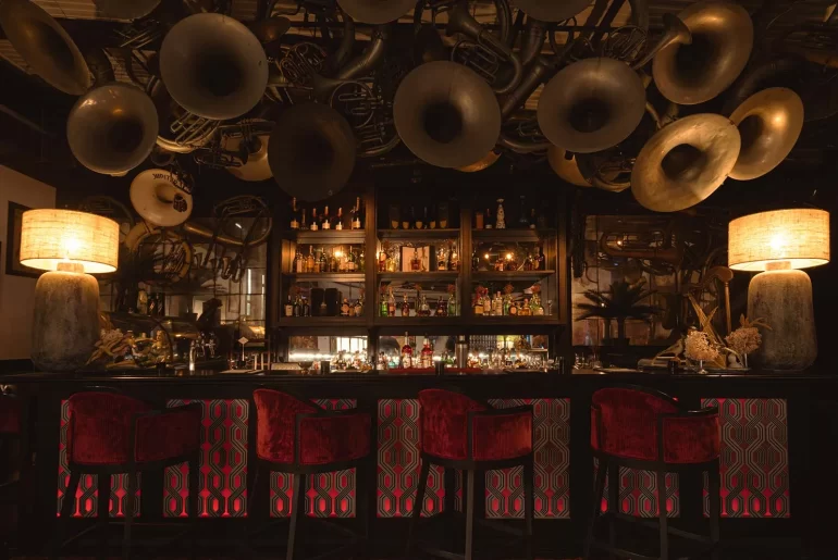 Interior of Thailand cocktail bar in Bangkok, with vintage brass instruments covering the ceiling above a backlit bar and red velvet stools — one of Bangkok's most distinctive luxury nightlife experiences
