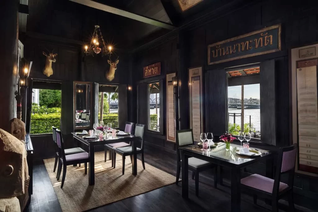 Chon Restaurant at The Siam Hotel Bangkok — an intimate Thai dining room inside a dark teak heritage house overlooking the Chao Phraya River, with antique signage, antler mounts, and candlelit tables set for dinner
