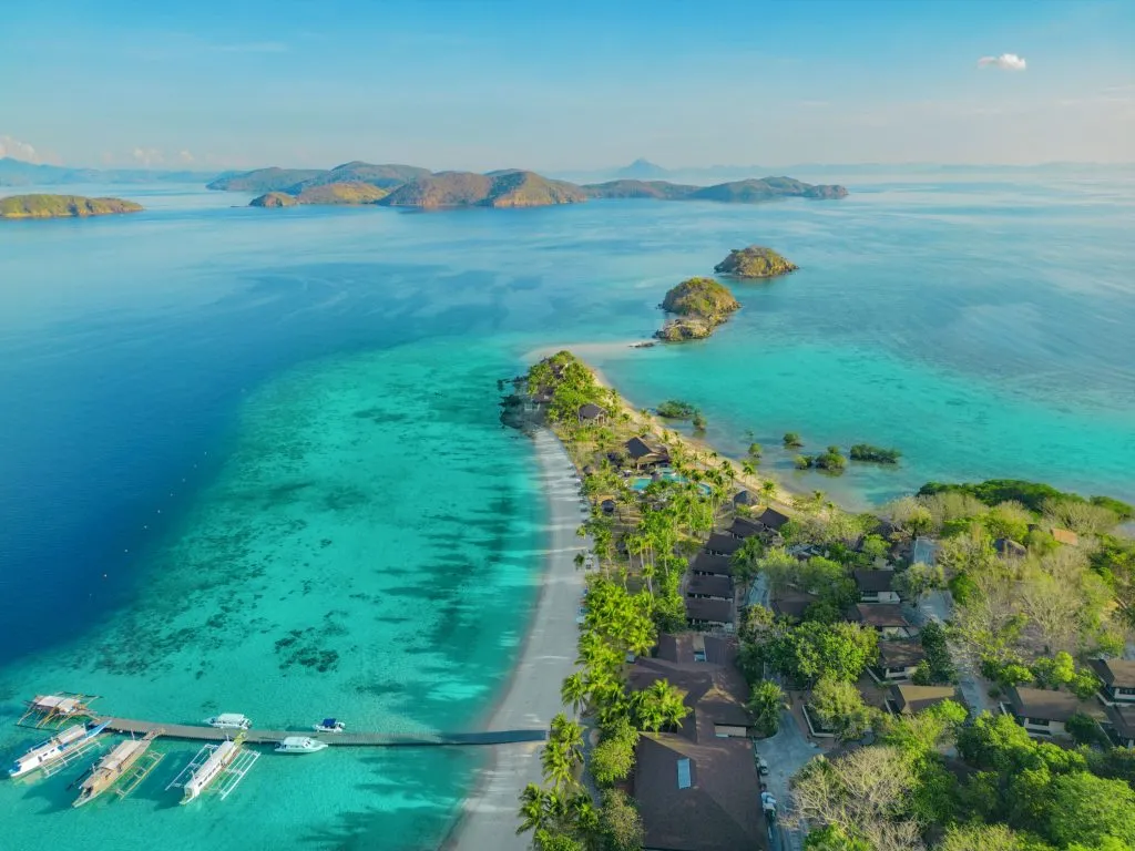 Aerial view of a luxury private island resort on a narrow sandbar surrounded by turquoise water and coral reefs in Coron, Palawan